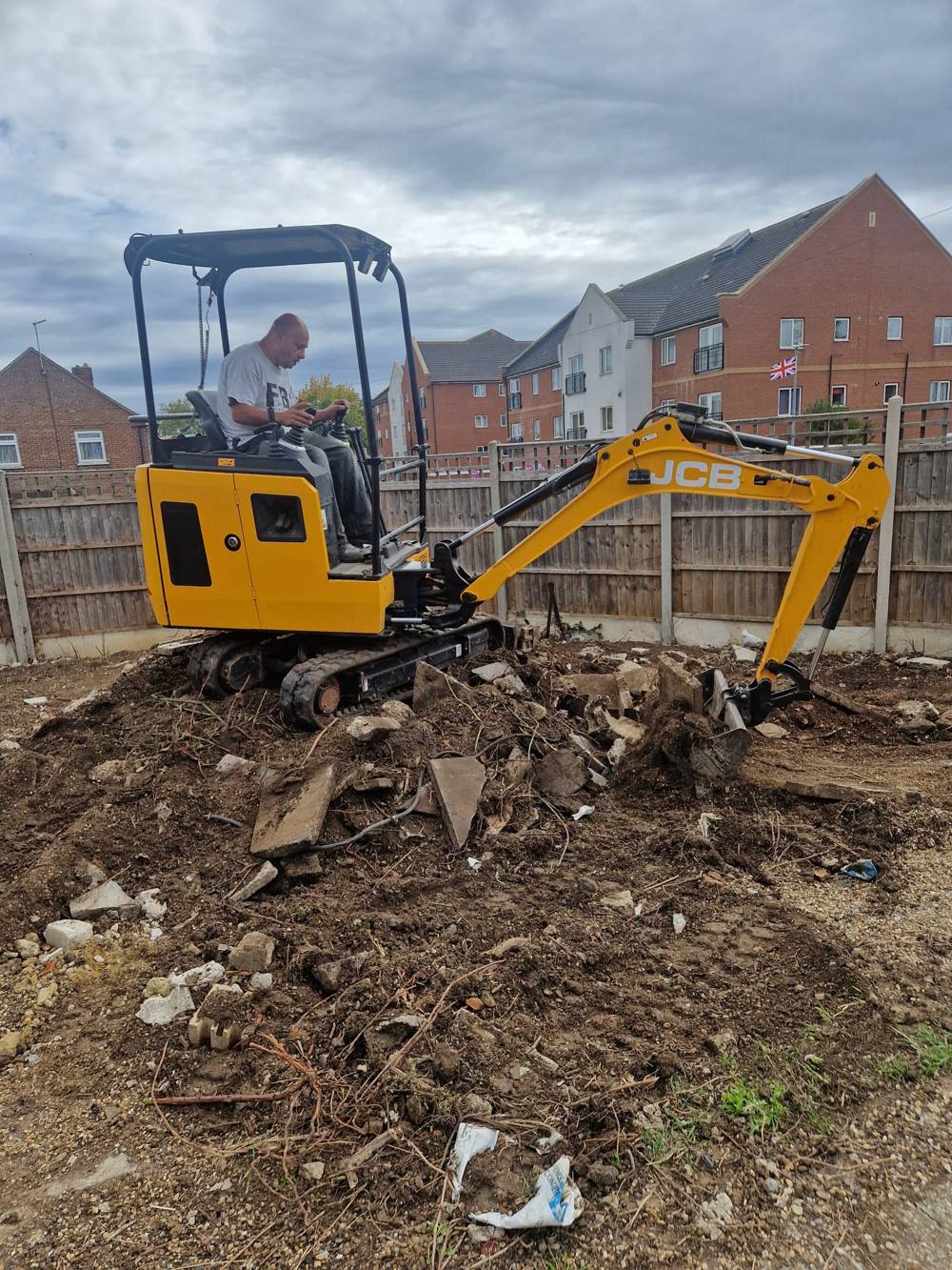 Yellow JCB mini digger with operator clearing rubble and levelling a garden plot