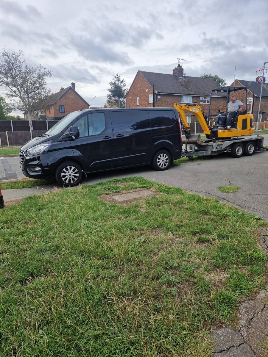 Mini digger on a plant trailer behind a black van, arriving at a job